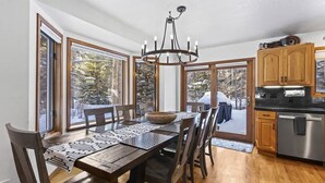 Un comedor con una mesa de madera, sillas y una araña. Se ven grandes ventanas con vistas a árboles.