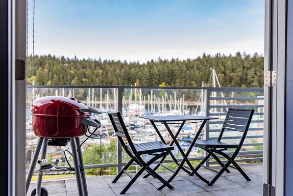 Een balkon met een barbecue, twee stoelen en uitzicht op boten en bomen.