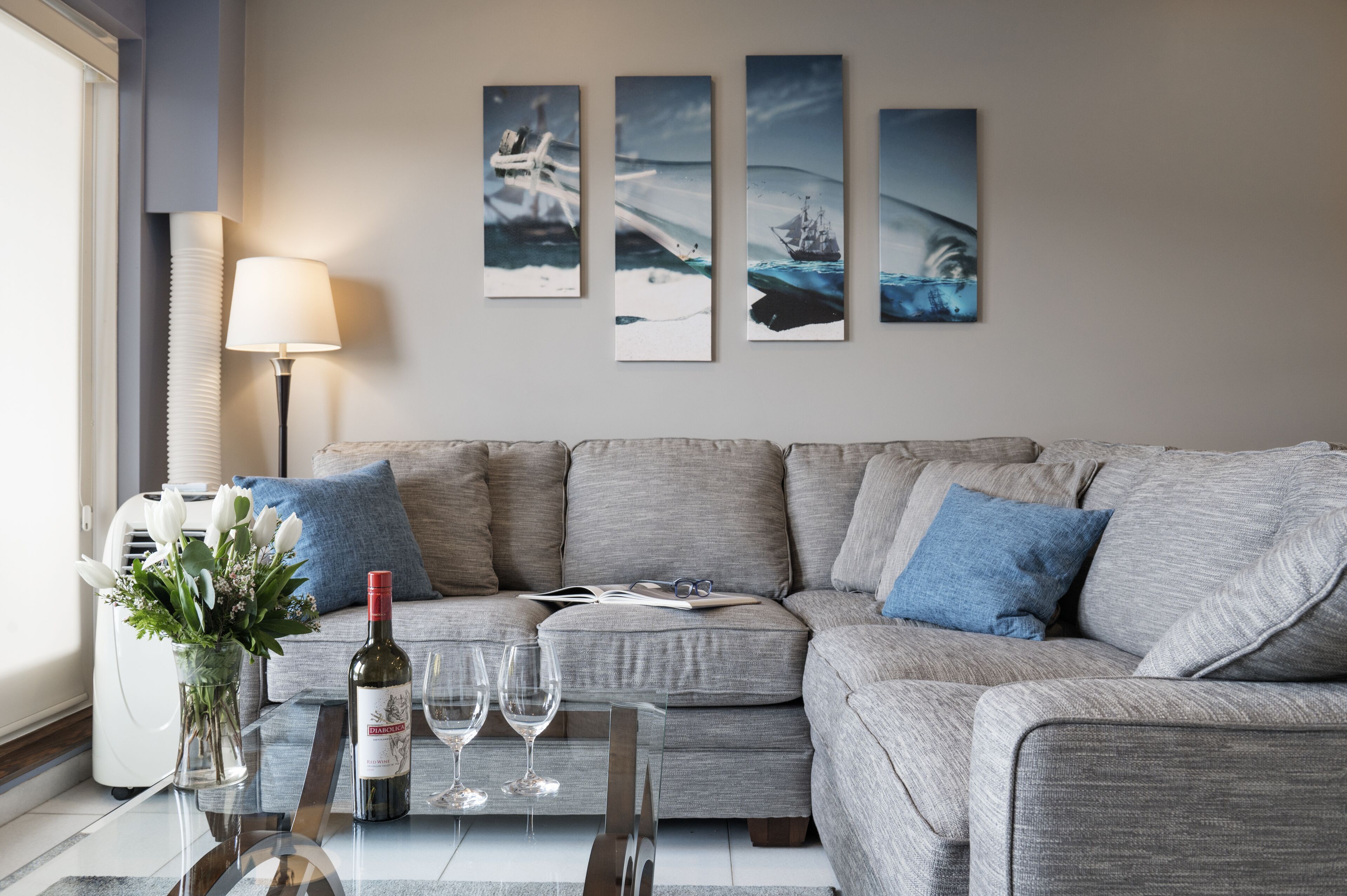 A living room with a grey sofa, a glass coffee table, a vase with flowers, a bottle of wine, and wine glasses. There are four framed pictures on the wall.