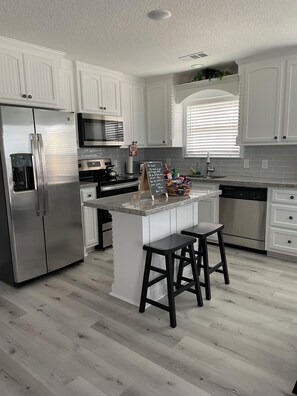 A modern kitchen with white cabinetry, stainless appliances, a central island with stools, and a chalkboard with a message. - Come and enjoy the beautiful Surfside beach and “heav’n fun” in this cozy home! (Freeport)