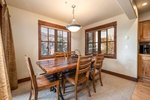 Un comedor con mesa y sillas de madera, ventanas amplias con persianas y una lámpara colgante.