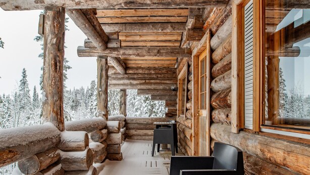 Een vakantiehuisje van hout met een houten veranda en uitzicht op besneeuwde bomen.