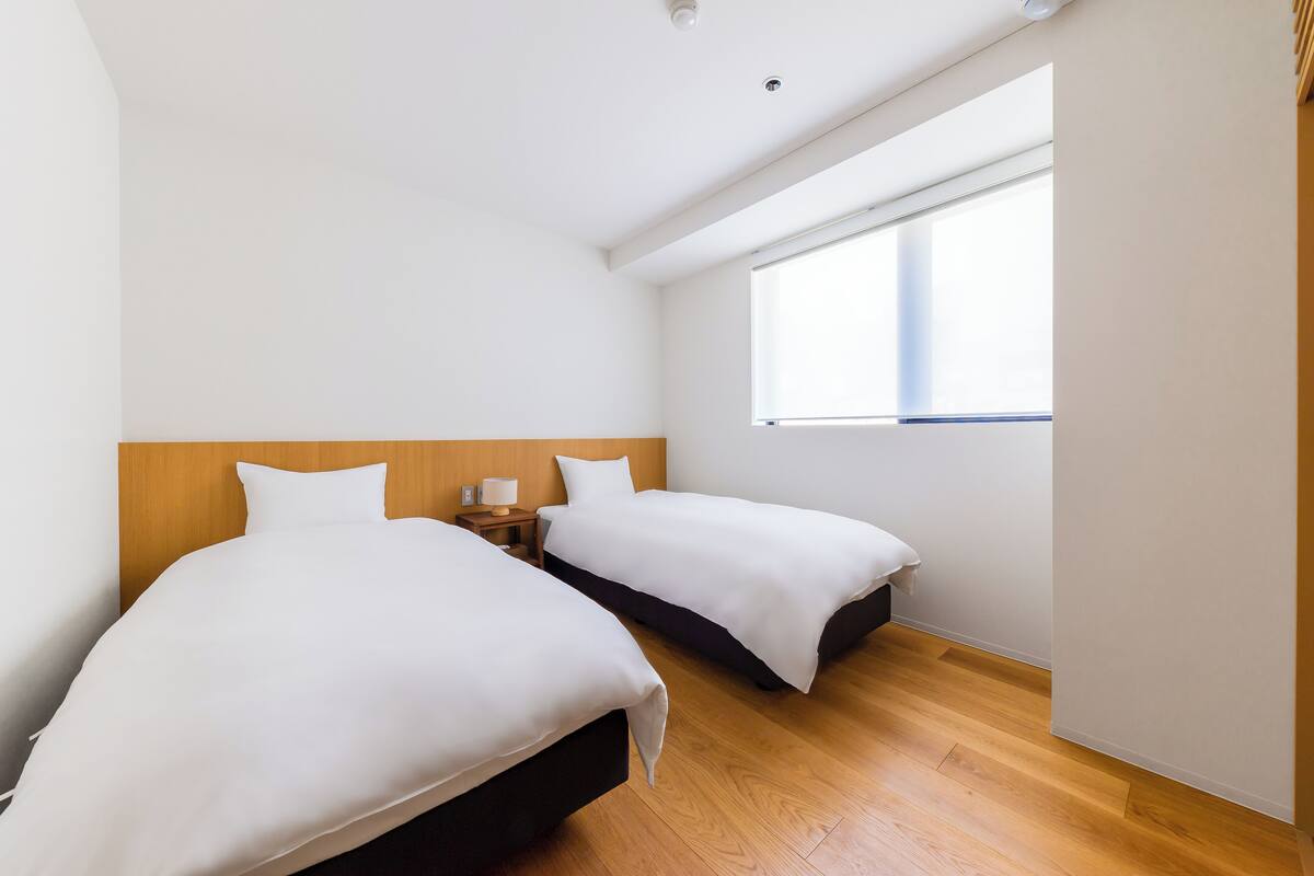 A hotel room with two beds, a wooden headboard, a nightstand with a lamp, and a large window providing natural light.