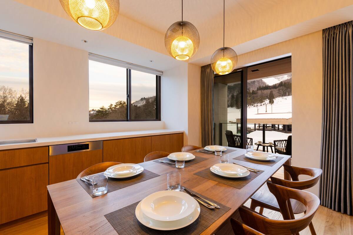 A modern dining area with a wooden table set for four, large windows, and hanging pendant lights.