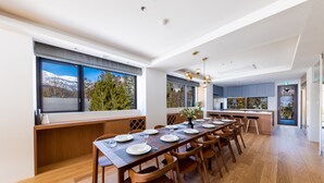 Un espace de repas moderne avec une longue table en bois pouvant accueillir six personnes, de grandes fenêtres offrant une vue sur les arbres et les montagnes, et une cuisine visible au fond.