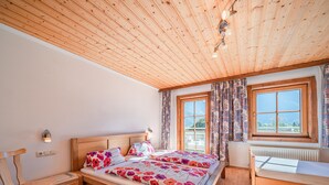 A bedroom with a wooden ceiling, a bed with floral bedding, a wooden nightstand, a chair, and a window with curtains.