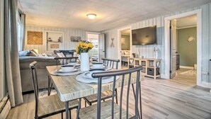A dining area with a wooden table set for four, surrounded by chairs. There is a vase with flowers on the table, and a television mounted on the wall. - Coastal Comfort Near the Bay! 'Buttermilk Cottage' (Wareham)