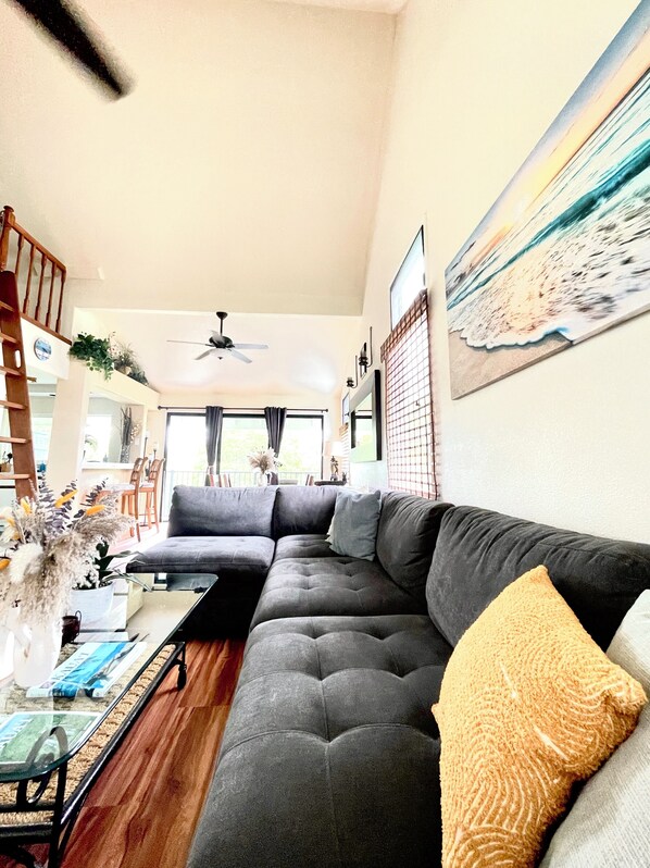 A living room with a sectional sofa, a ceiling fan, a dining area with chairs, and a painting on the wall.
