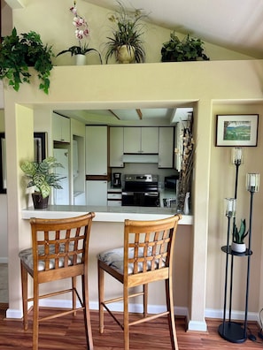A dining area with two wooden chairs, a kitchen in the background, and potted plants on the walls. - Elegant Princeville Condo with Cozy Loft Bedroom and Mountain Views (Princeville)