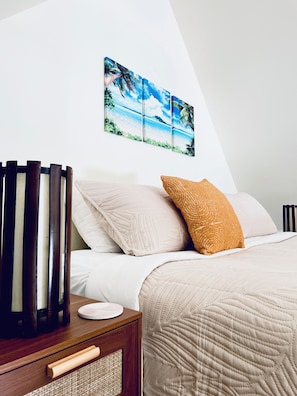 Un lit bien fait, recouvert d’une courtepointe, une table de chevet en bois avec une lampe, et un tableau représentant une plage tropicale accroché au mur.