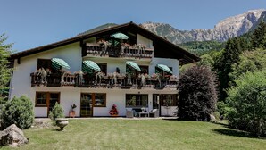 A two-story building with balconies adorned with flowers and green umbrellas, surrounded by a well-maintained lawn and trees, with mountains in the background. - Vacation apartment Untersberg 2 persons, 52 sqm (Schönau am Königssee)