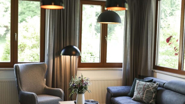Una sala de estar moderna con sofá, sillón, mesa de comedor con alimentos para el desayuno y ventanas grandes con cortinas.