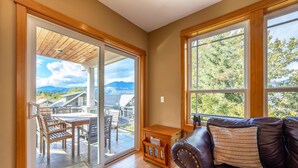 A room with a sliding glass door leading to a patio with a table and chairs, a brown leather sofa with cushions, and large windows with a view of greenery and mountains.