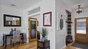 Un couloir doté d’un plancher en bois, d’un miroir, d’une chaise avec un coussin à motifs, d’une petite table sur laquelle se trouve un vase, et d’une porte qui donne sur une autre pièce.