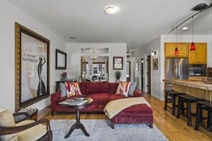 A spacious living room with a red sofa, a coffee table, and a kitchen area with wooden cabinets and a stainless steel refrigerator.