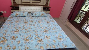 Ein Schlafzimmer mit floralem Bettüberwurf, einem Holzkopfende, einem Sessel und einem fenster mit Vorhängen.