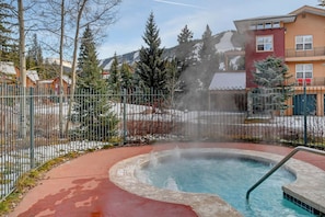 Un bain à remous entouré d’une clôture métallique, avec une montagne en arrière-plan et un bâtiment doté d’un toit rouge.
