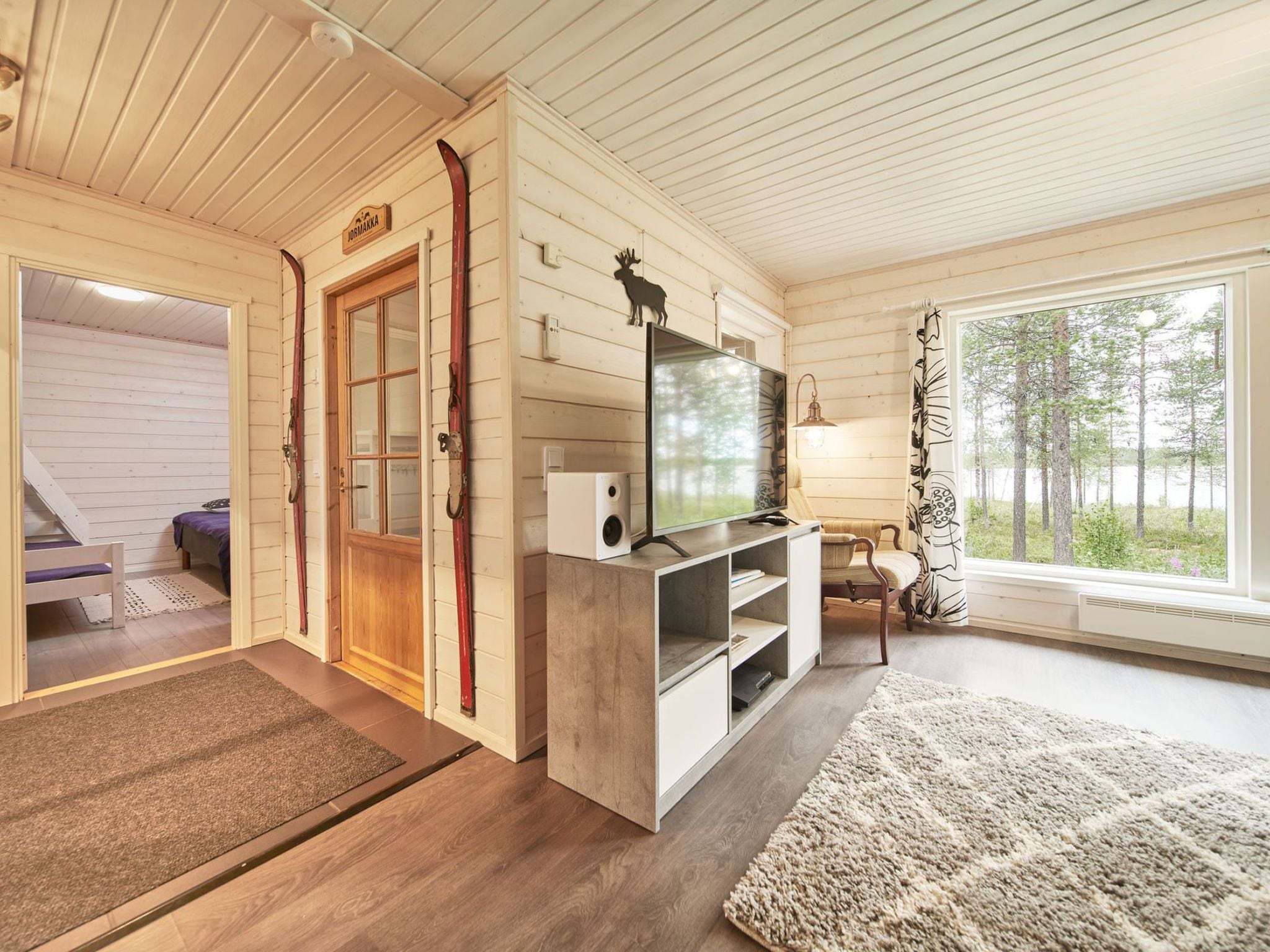 A cozy cabin interior with a wooden interior, a television on a stand, a sofa, and a window with a view of trees.