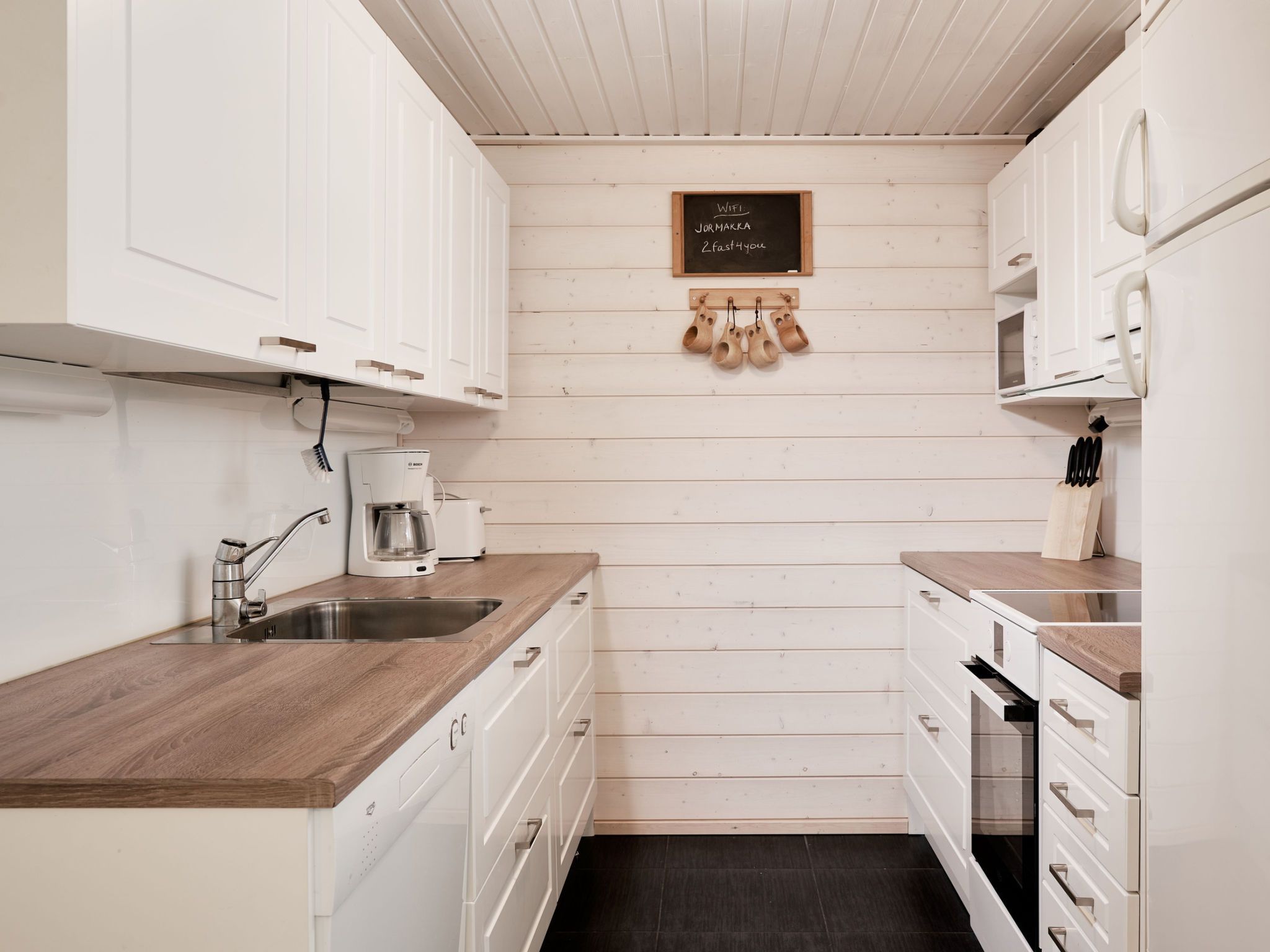 A compact kitchen with white cabinetry, a wooden countertop, a coffee maker, and a chalkboard sign.