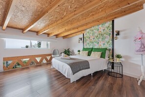 Une chambre avec un plafond en bois, un lit recouvert d’une courtepointe blanche, un oreiller vert, une petite table ronde avec une lampe et une fenêtre donnant sur un beau paysage.