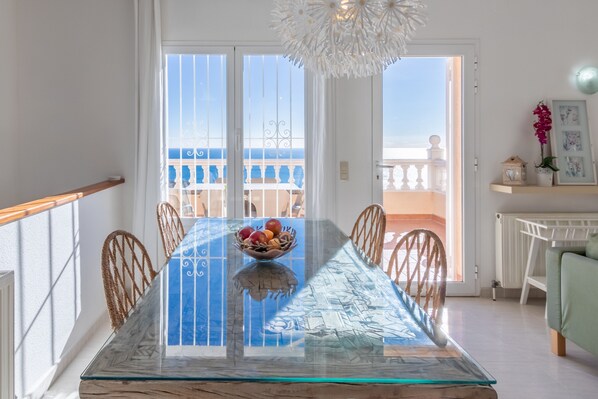 A dining table with a glass top and a bowl of fruit, a white railing, and a view of the sea through a balcony door.