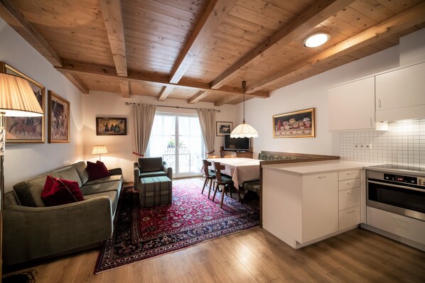 Eine moderne Küche mit weißen Schränken, ein Essbereich mit Tisch und Stühlen, ein Sofa mit roten Kissen und ein gemustertes Teppich.