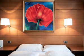 Et hotelværelse med en seng, to sengeborde, en vægmonteret lampe og et indrammet billede af en rød blomst.