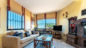 A living room with a sofa, coffee table, TV, fireplace, and large windows. - Villa 'Cosy House Jacuzzi Wifi Pool View' with Sea View, Wi-Fi and Air Conditioning (Mijas Pueblo)