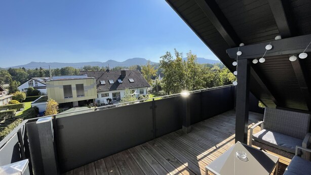 Une terrasse en bois avec des meubles d’extérieur, une vue sur les maisons et les montagnes, et des guirlandes lumineuses.