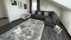 Un salon moderne avec un canapé gris, une table d’appoint blanche, un tapis à poils longs gris et une fenêtre avec des rideaux.