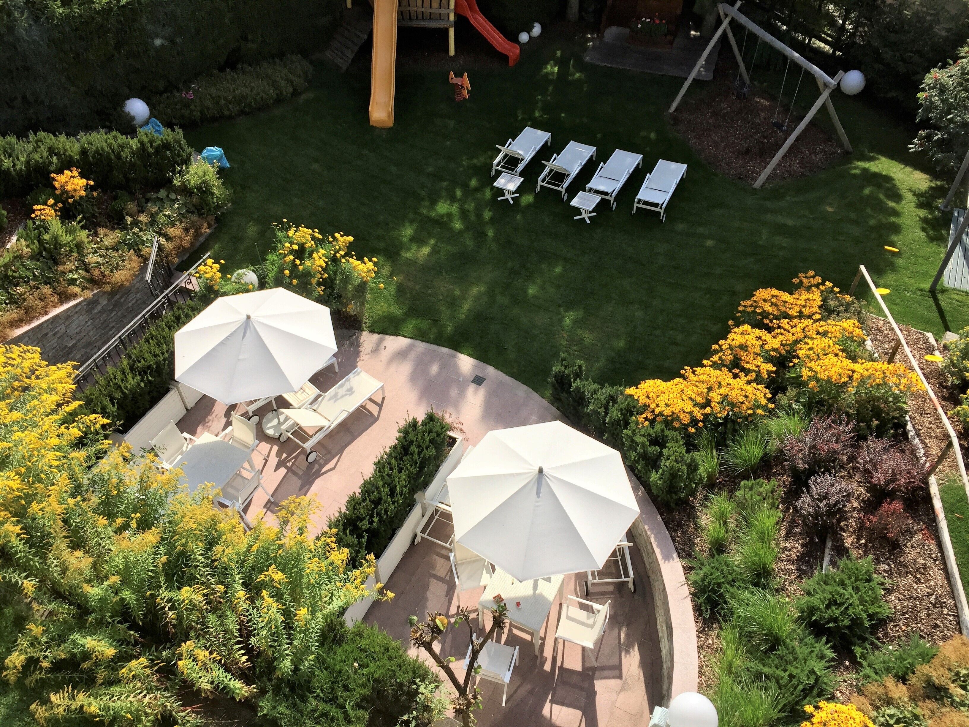 En terrasse med udendørs møbler og parasoller, omgivet af anlagte haver og en legeplads.
