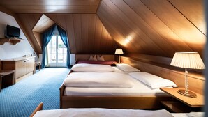Ein Zimmer mit schrägen Wänden, Holzwänden, einem Bett mit weißer Bettwäsche, einem Nachttisch mit Lampe, einem an der Wand montierten Fernseher und einem Fenster mit Vorhängen.