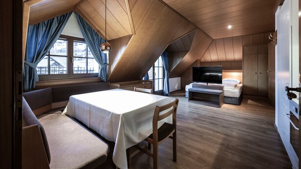 Un dormitorio tipo loft moderno con techo inclinado, una cama grande, una zona de estar con mesa y un televisor en una esquina.