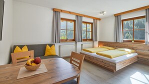 Quarto com cama de madeira, um sofá, mesa de jantar com frutas e janelas amplas com cortinas.