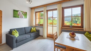 Een moderne kamer met een grijze bank, een houten eettafel en grote ramen met uitzicht op groen en gebouwen.