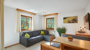 Un salon moderne avec un canapé d’angle gris, une table à manger en bois et de grandes fenêtres avec des rideaux.