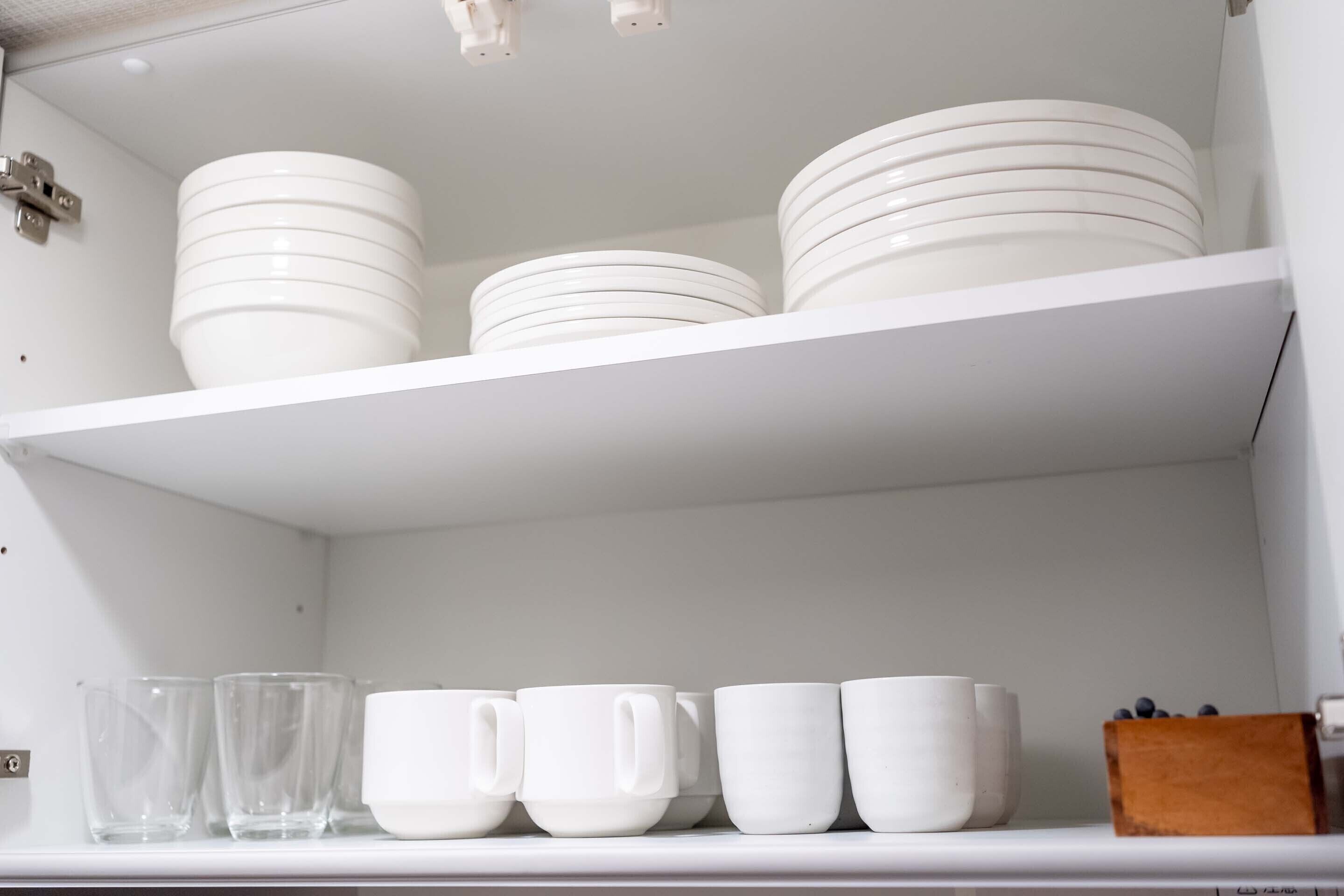 Una estantería blanca con platos y cuencos blancos apilados, y una fila de tazas blancas debajo.