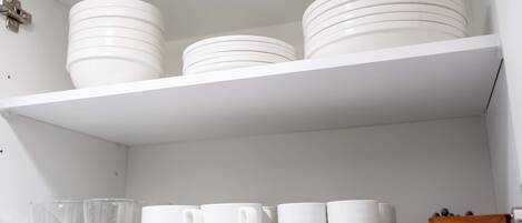 Una estantería blanca con platos y cuencos blancos apilados, y una fila de tazas blancas debajo.