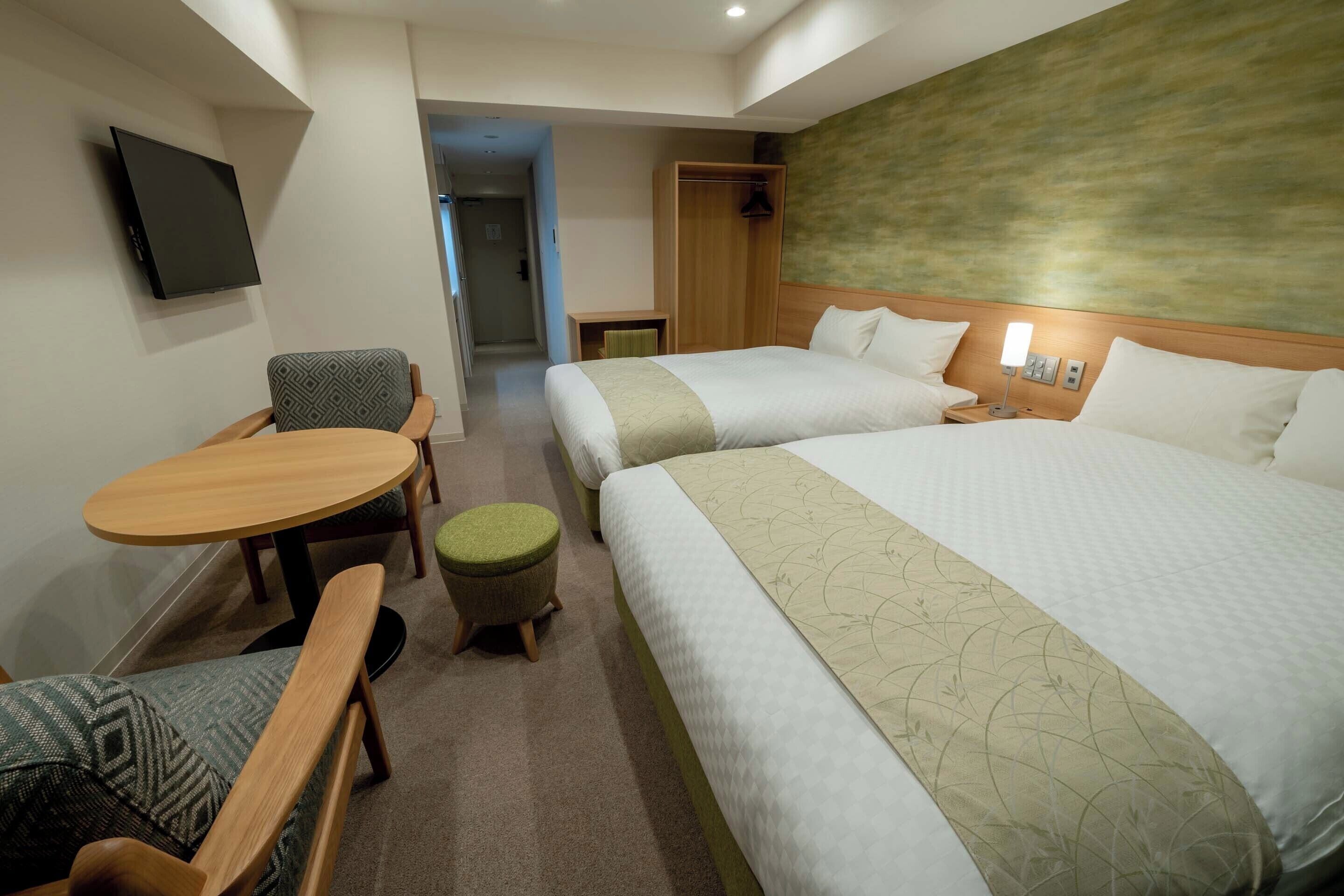 A hotel room with two beds, a TV mounted on the wall, a wooden table, and a small stool.