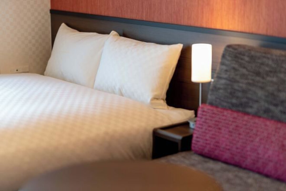 Quarto de hotel com uma cama bem arrumada, uma luminária de cabeceira e uma pequena mesa de apoio.