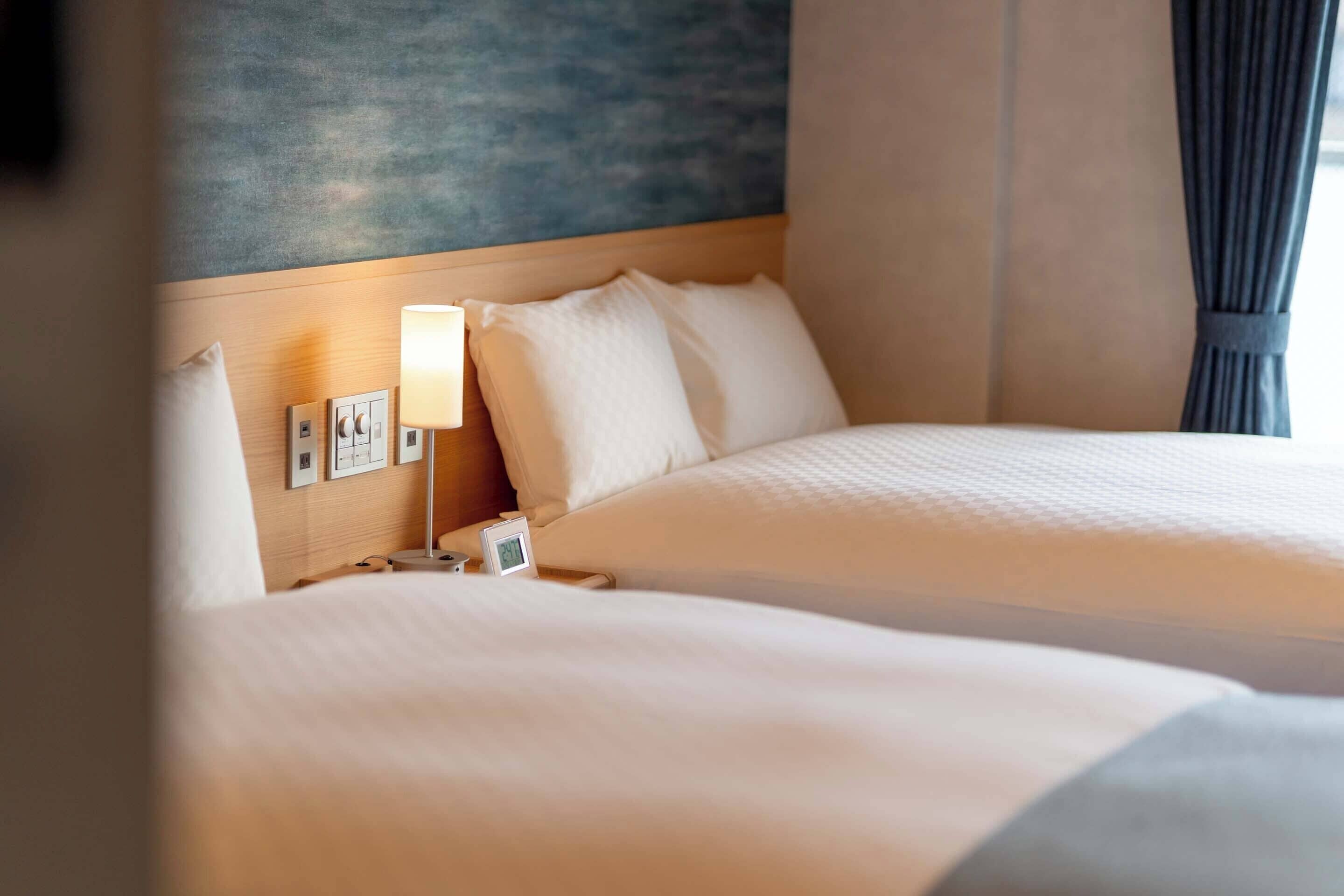 A hotel room with two beds, a nightstand with a lamp, a clock, and a wall-mounted control panel.