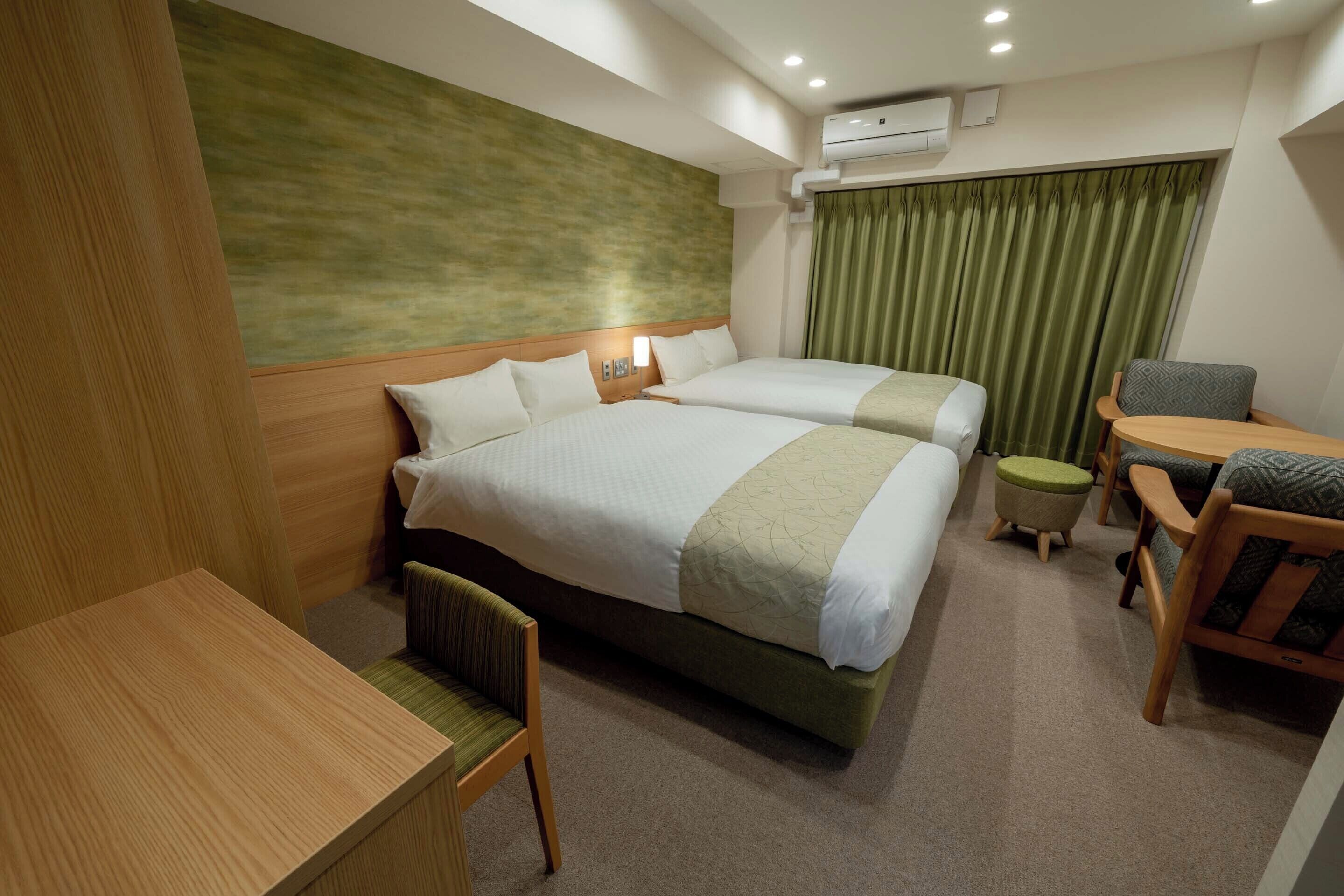 A hotel room with two beds, a desk, a chair, a small table, and a green curtain.