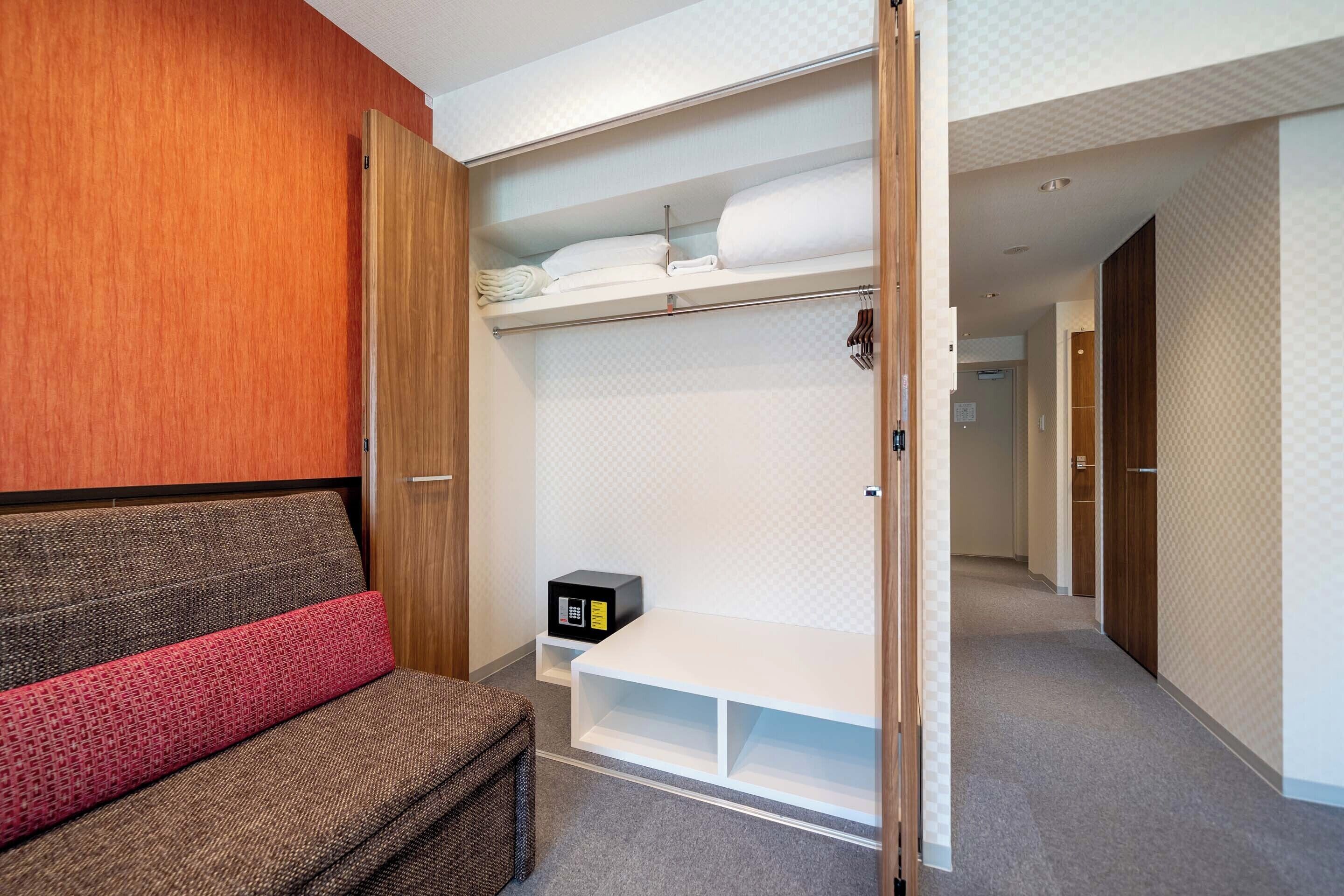 A hotel room with a bed, a bench, a wardrobe, and a small table with a black object on it.