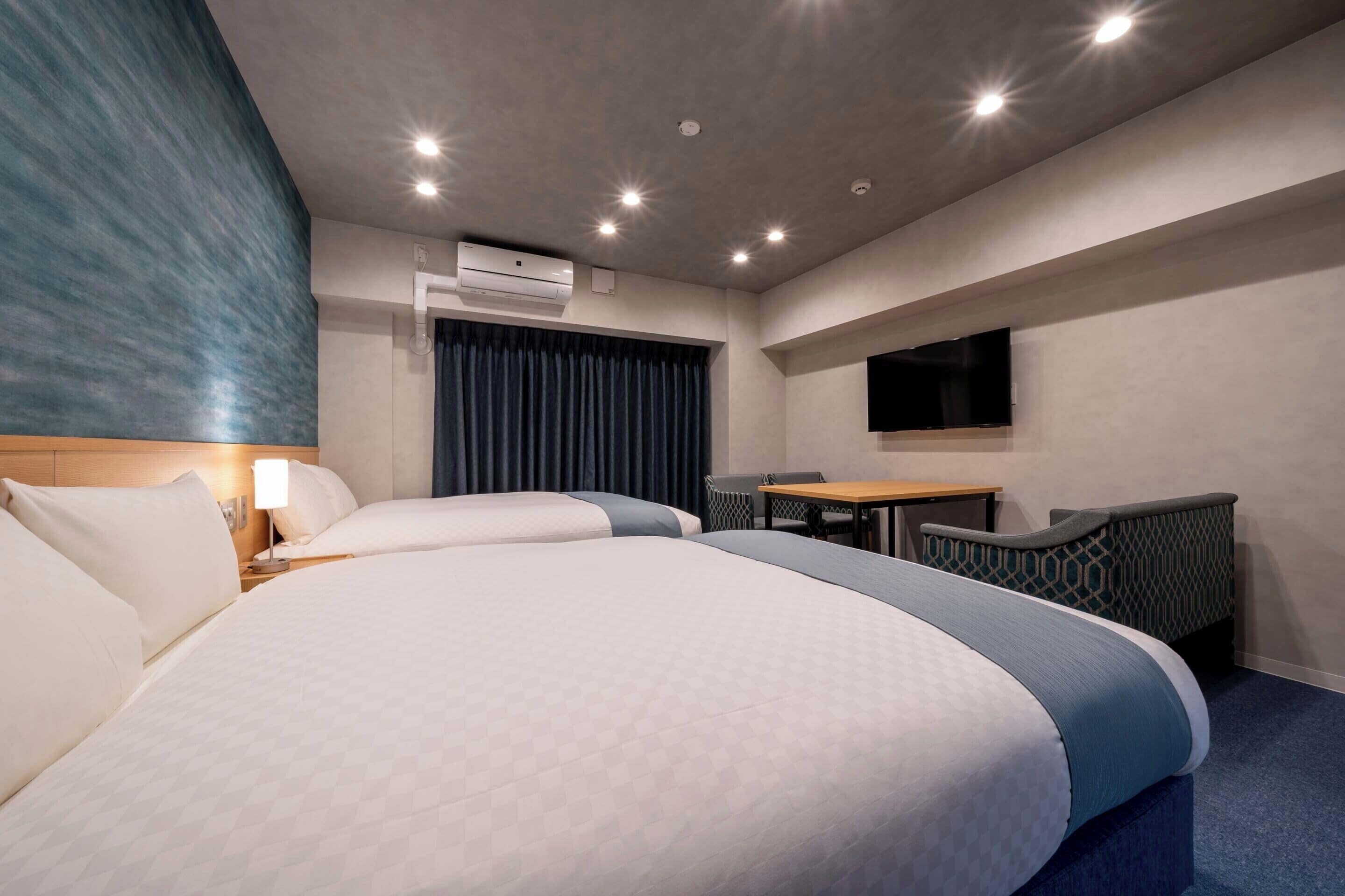 A hotel room with two beds, a desk, a chair, an air conditioning unit, and a flat-screen TV mounted on the wall.