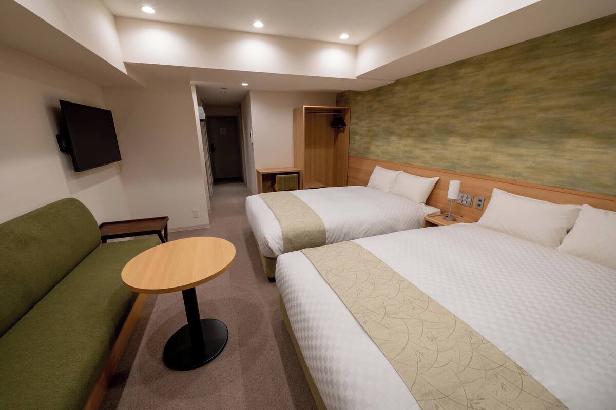 Ein Hotelzimmer mit zwei Betten, einem grünen Sofa, einem runden Holztisch und einem an der Wand montierten Fernseher.