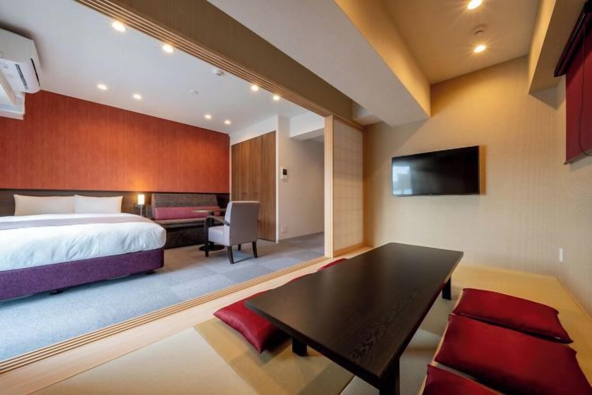 A modern hotel room with a large bed, a desk, a TV, and a dining table.