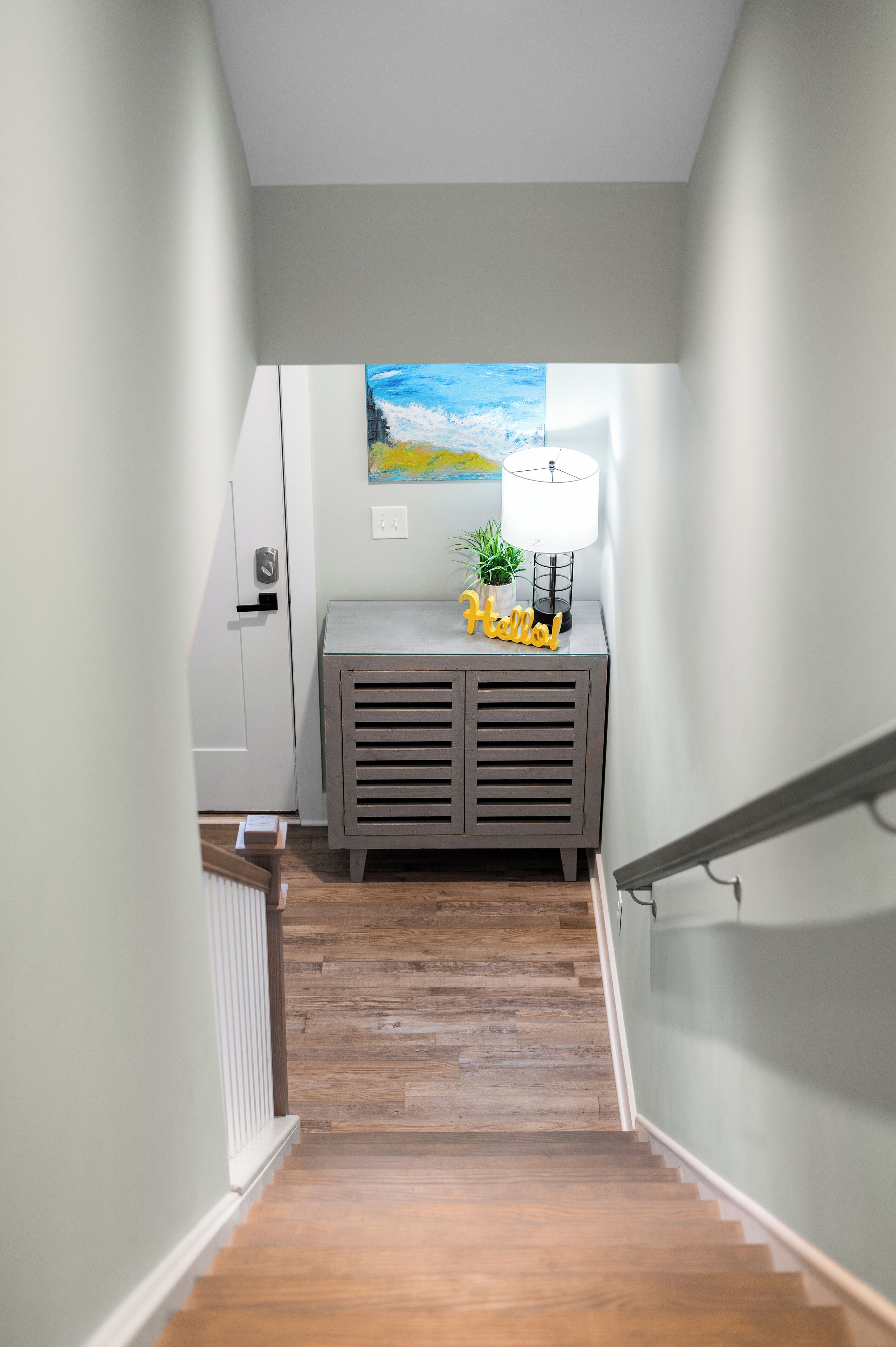 A narrow hallway with wooden flooring, a gray console table with a lamp and a small plant, and a painting on the wall.