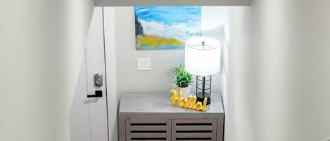 A narrow hallway with wooden flooring, a gray console table with a lamp and a small plant, and a painting on the wall.