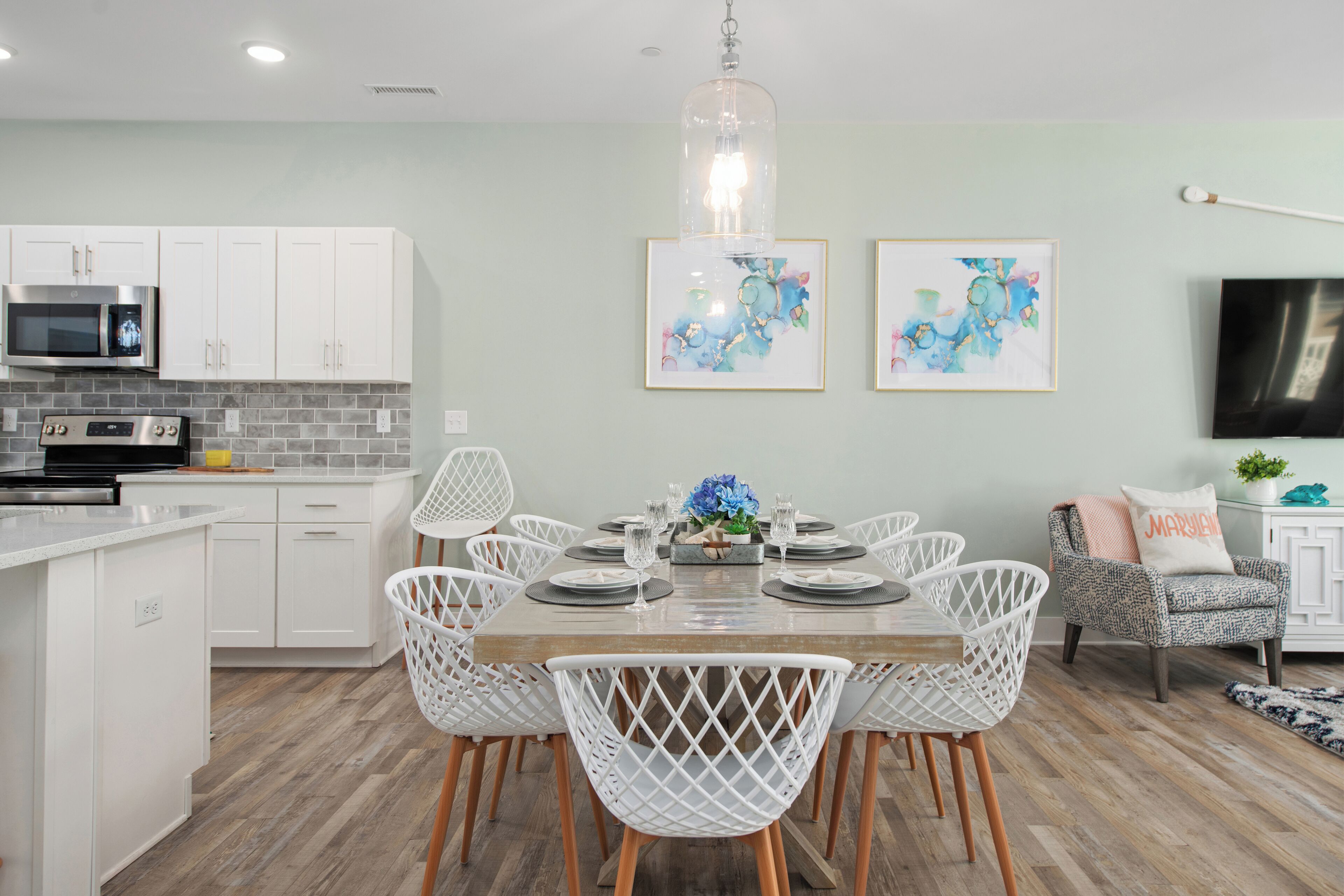 Una cocina moderna con gabinetes blancos, microondas y un comedor con mesa de madera y sillas blancas. Hay dos obras de arte enmarcadas en la pared y un televisor a la derecha.