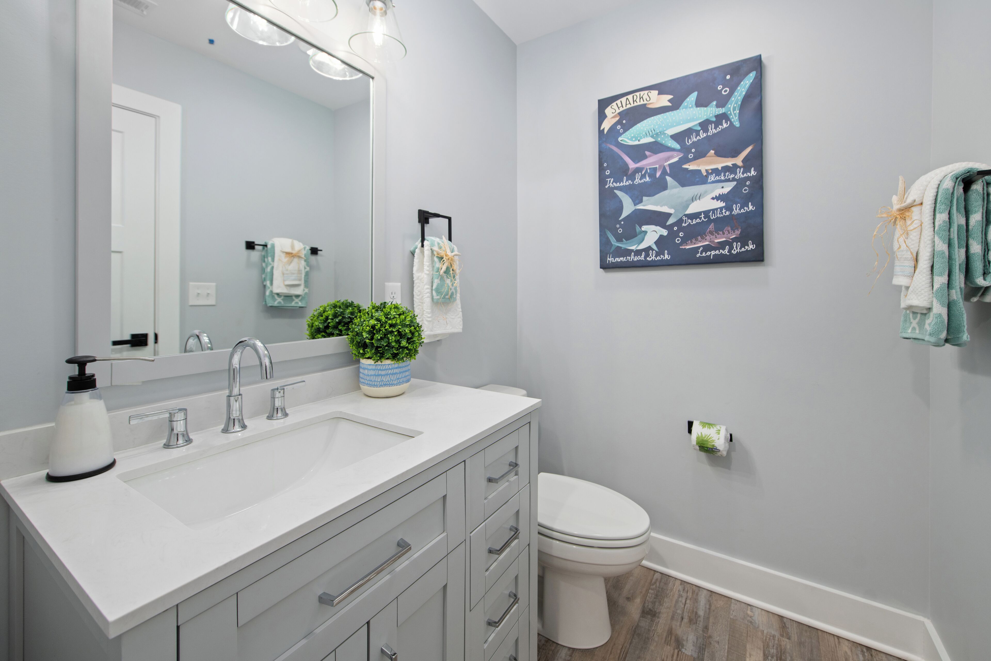 A modern bathroom with a white sink, two faucets, a mirror, and a toilet. There is a wall-mounted towel rack with towels and a framed shark-themed artwork.